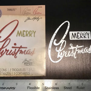 May include: A metal die set for crafting, featuring the words "Merry Christmas" in a stylized script font. The die set is designed to be used with a die-cutting machine to create intricate designs for cards, scrapbook pages, and other paper crafts.
