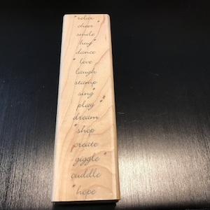 May include: A wooden block with the words "relax", "cheer", "smile", "hug", "dance", "love", "laugh", "stamp", "sing", "play", "dream", "shop", "create", "giggle", "cuddle", and "hope" engraved on it.