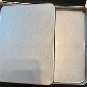 May include: A silver metal rectangular tin with a lid. The tin is empty and has a smooth, slightly textured surface.