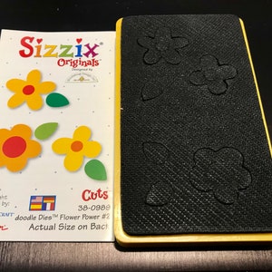 May include: A yellow and black die-cutting machine with a black cutting mat. The mat has a design of four flowers and four leaves. The machine is labeled "Sizzix Originals" and "Cuts 38-0989 doodle Dies™ Flower Power #2 Actual Size on Back".