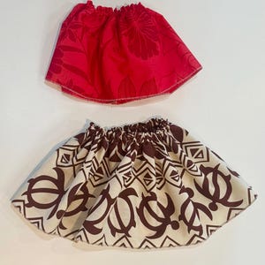 May include: Two small skirts. The top skirt is red with a floral pattern. The bottom skirt is off-white with a brown turtle and geometric pattern. Both skirts have elastic waistbands and a flared shape.
