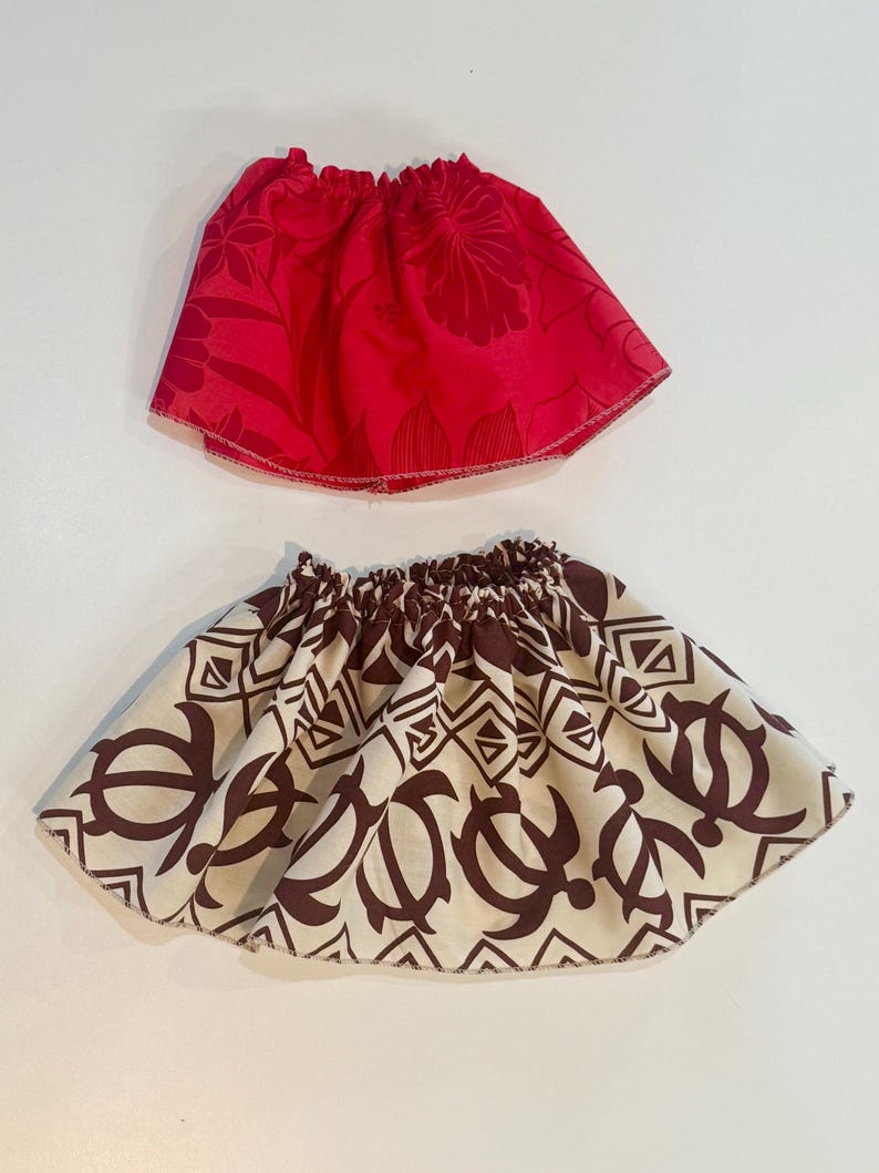 May include: Two small skirts. The top skirt is red with a floral pattern. The bottom skirt is off-white with a brown turtle and geometric pattern. Both skirts have elastic waistbands and a flared shape.