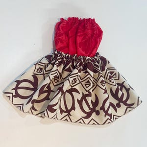 May include: A red and brown baby skirt with a Hawaiian design. The top is red with a floral pattern, and the skirt features a repeating pattern of brown turtles and geometric shapes on a cream background. The skirt is gathered at the waist.