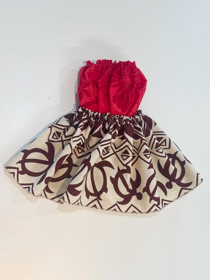May include: A red and brown baby skirt with a Hawaiian design. The top is red with a floral pattern, and the skirt features a repeating pattern of brown turtles and geometric shapes on a cream background. The skirt is gathered at the waist.