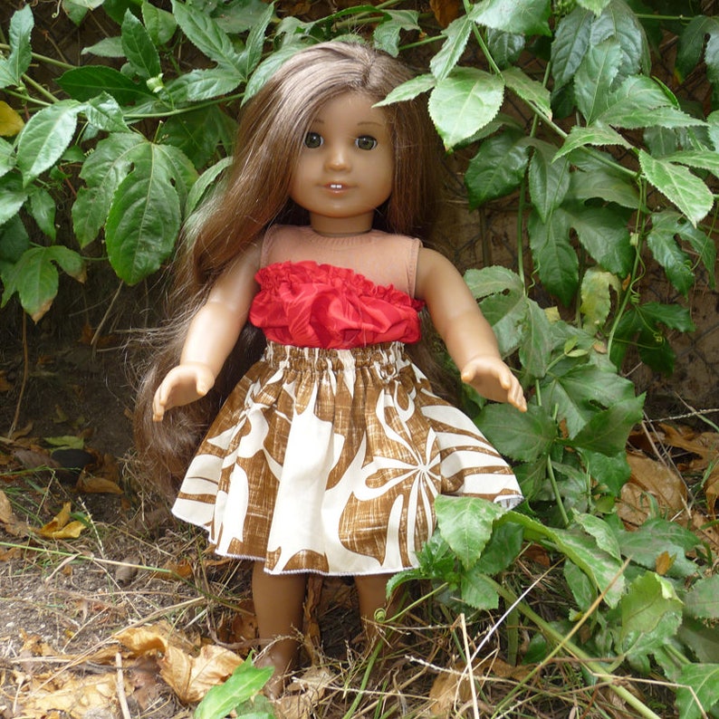 May include: A doll with long brown hair, wearing a tan top with a red ruffled accent and a brown and white patterned skirt. The doll is posed outdoors among green foliage.