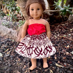 May include: A doll wearing a red top and a skirt with a white background and a brown turtle pattern. The doll has brown hair and is standing on a bed of rocks and dirt. The doll is outdoors, with green foliage in the background.