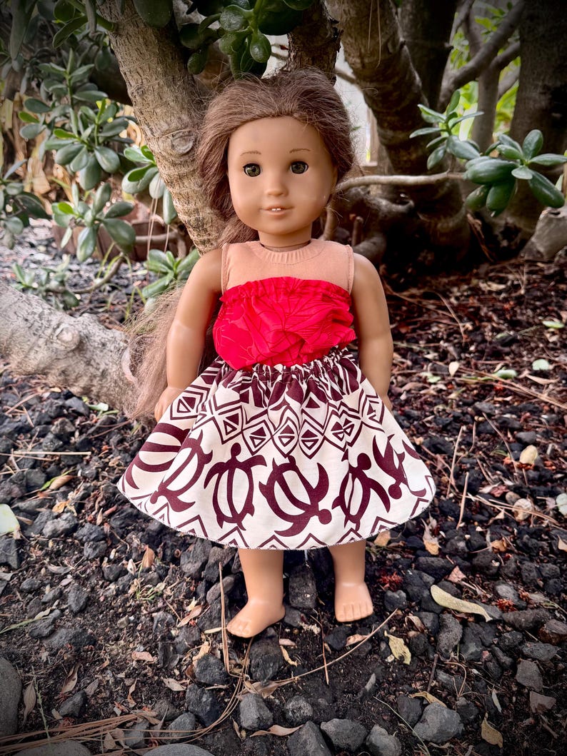 May include: A doll wearing a red top and a skirt with a white background and a brown turtle pattern. The doll has brown hair and is standing on a bed of rocks and dirt. The doll is outdoors, with green foliage in the background.