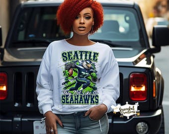 May include: White sweatshirt featuring the Seattle Seahawks logo and text. The graphic includes a football player and the words "They Not Like Us Seahawks." The sweatshirt is worn by a person in light blue jeans.