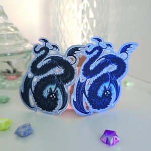 May include: Two stickers featuring a stylized bat design. One sticker is black with a light blue outline, and the other is a vibrant blue with a darker blue outline. The bats have detailed wings and a glowing center.