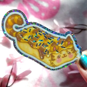 May include: A die-cut sticker featuring a cat shaped like a long pastry with a curled tail. The cat has light brown fur, pink cheeks, and is covered in brown icing with blue sprinkles. The sticker has a reflective, holographic glitter border.