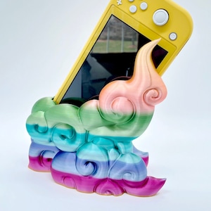 May include: A yellow Nintendo Switch console is held in a 3D printed stand shaped like a rainbow cloud. The stand is made of multiple layers of different colors, including pink, green, blue, and purple.
