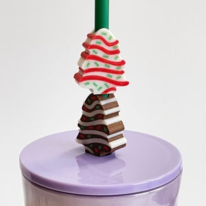 May include: A purple reusable cup with a lid and a straw. The straw has two Christmas tree-shaped charms, one red and white and one brown and white.