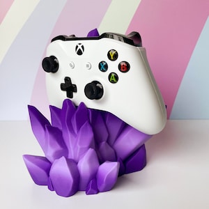 May include: A white Xbox controller with black buttons and a purple crystal-shaped stand. The controller has the letters A, B, X, and Y on the buttons.
