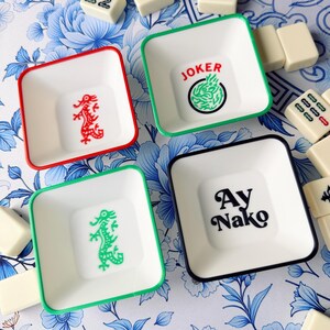 May include: Four square white dishes with colored rims and designs. One dish has a red rim and a red dragon, another has a green rim and a green dragon, a third has a green rim with the word "JOKER" and a green design, and the last has a black rim with the words "Ay Nako".
