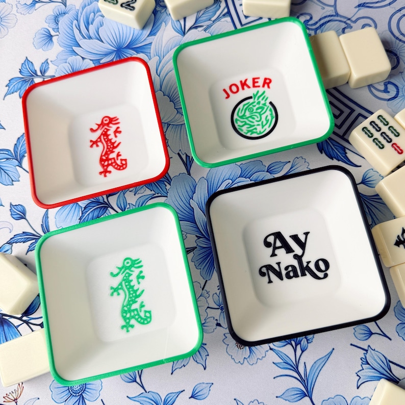 May include: Four square white dishes with colored rims and designs. One dish has a red rim and a red dragon, another has a green rim and a green dragon, a third has a green rim with the word "JOKER" and a green design, and the last has a black rim with the words "Ay Nako".