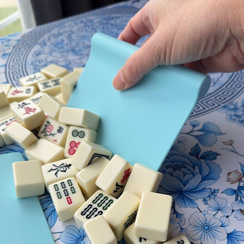 K&ouml;nnte beinhalten: Ein hellblauer Mahjong-Kartenhalter mit gebogenem Rand wird verwendet, um elfenbeinfarbene Spielsteine zu sammeln. Die Spielsteine haben rote, gr&uuml;ne und schwarze Symbole. Das Spiel befindet sich auf einer blau-wei&szlig;en Oberfl&auml;che mit Blumenmuster.