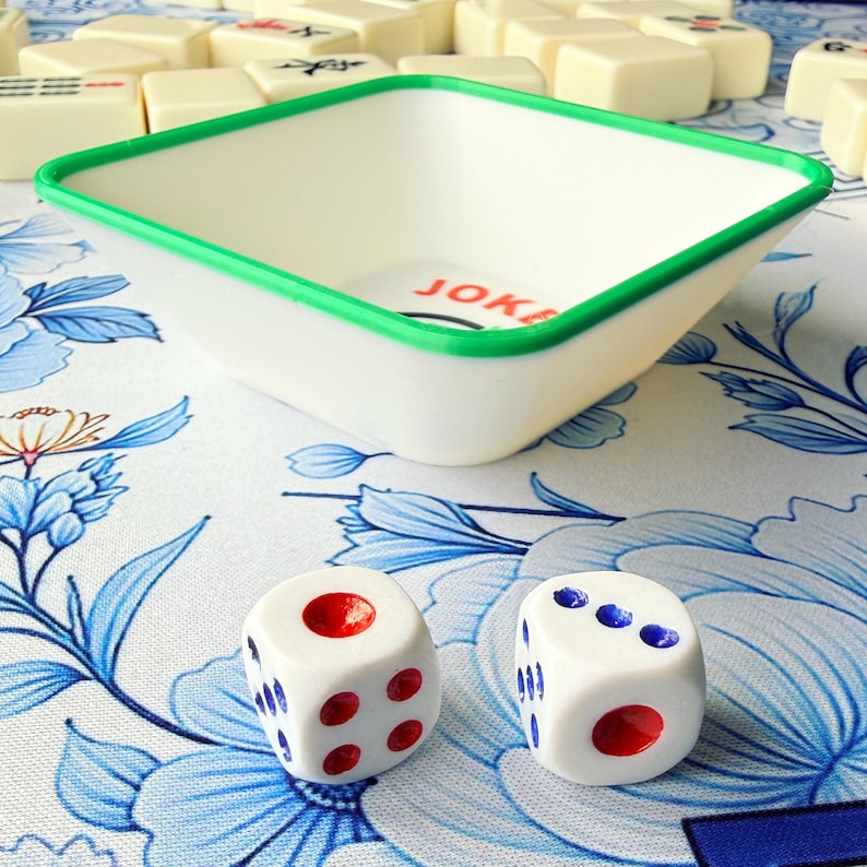 May include: A white and green Mahjong game tray with the word "JOKE" printed inside. Two white dice with red and blue dots are in the foreground. The tray and dice are on a floral patterned surface. Other Mahjong tiles are in the background.