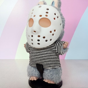 May include: A plush toy bunny wearing a white hockey mask and a black and white striped shirt. The bunny has gray fur and is standing on a black base. The background is a pastel rainbow.