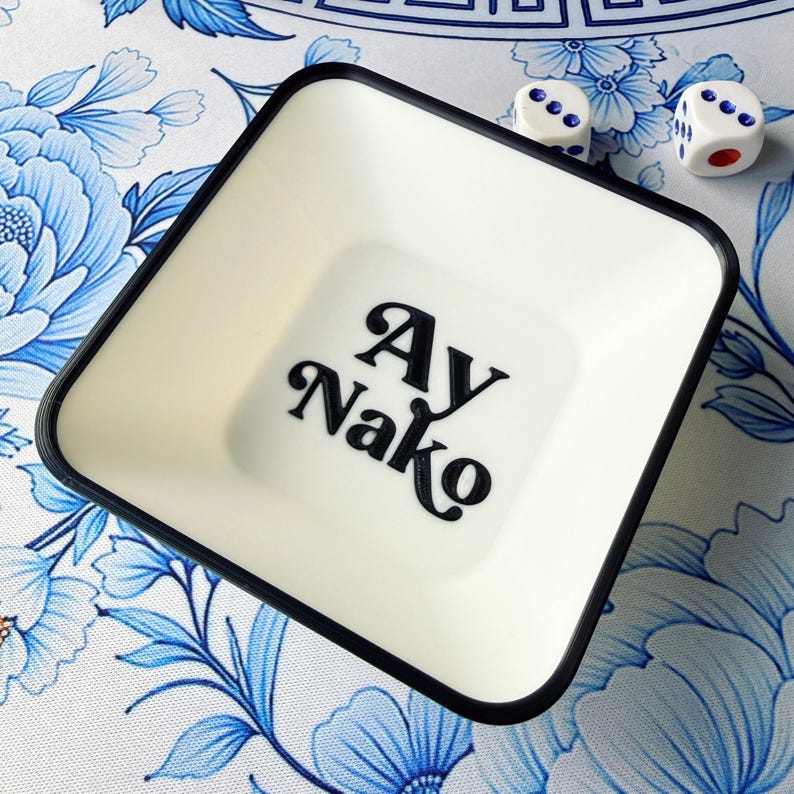 May include: A square, off-white dish with a black rim and the text "Ay Nako" in black script. Two white dice with blue and red dots are next to the dish. The dish is on a floral patterned surface with blue flowers.