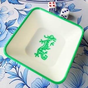 May include: A square, off-white tray with a green border and a green dragon design in the center. Two white dice with red and blue dots are next to the tray. The tray sits on a blue and white floral patterned surface.