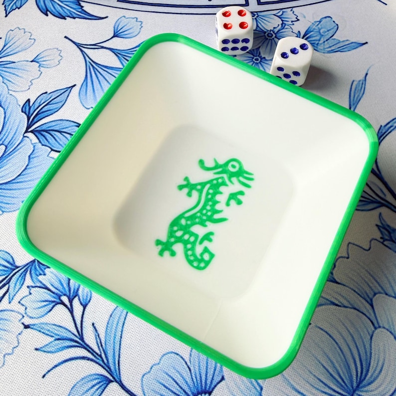 May include: A square, off-white tray with a green border and a green dragon design in the center. Two white dice with red and blue dots are next to the tray. The tray sits on a blue and white floral patterned surface.