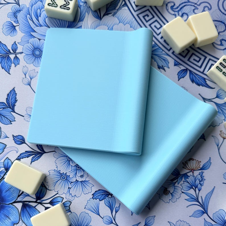 K&ouml;nnte beinhalten: Zwei hellblaue Mahjong-Kartenhalter werden auf einer Oberfl&auml;che mit floralem Muster gezeigt. Die Halter sind rechteckig mit einer gebogenen Kante. Mehrere Mahjong-Karten sind um die Halter herum verstreut.