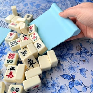 K&ouml;nnte beinhalten: Elfenbeinfarbene Mahjong-Steine mit roten und schwarzen Symbolen werden aus einem hellblauen Tablett gegossen. Die Steine sind auf einer blau-wei&szlig; gemusterten Oberfl&auml;che verstreut. Die Spielsteine sind rechteckig.