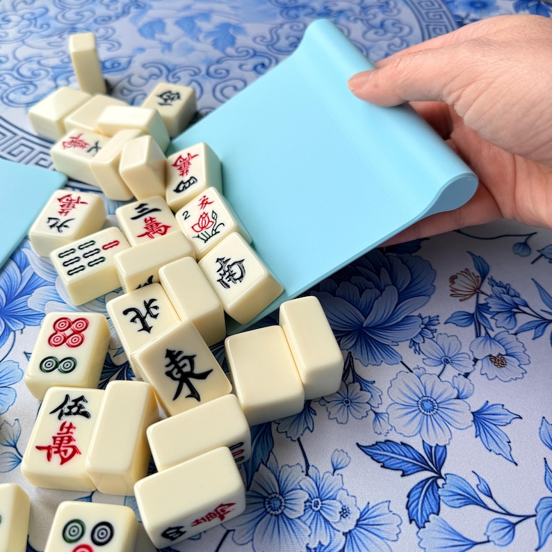 K&ouml;nnte beinhalten: Elfenbeinfarbene Mahjong-Steine mit roten und schwarzen Symbolen werden aus einem hellblauen Tablett gegossen. Die Steine sind auf einer blau-wei&szlig; gemusterten Oberfl&auml;che verstreut. Die Spielsteine sind rechteckig.