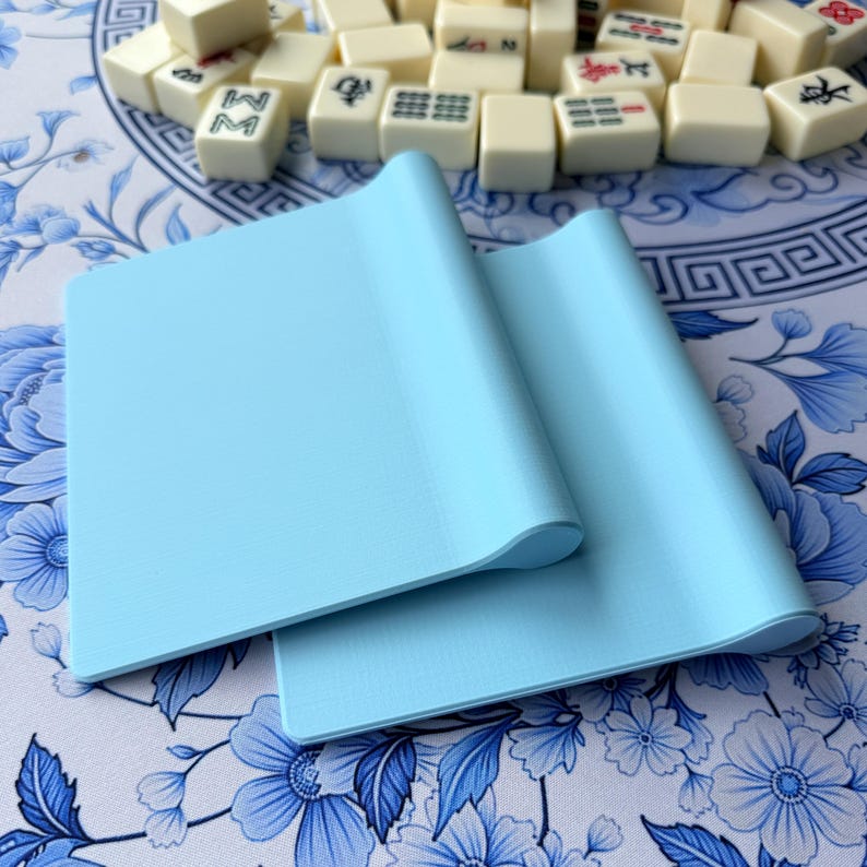 K&ouml;nnte beinhalten: Zwei hellblaue, quadratische Mahjong-Kartenhalter werden auf einer blau-wei&szlig; gemusterten Oberfl&auml;che mit Blumenmuster gezeigt. Die Halter sind aus weichem Material und haben eine gebogene Kante. Mahjong-Steine sind im Hintergrund verstreut.