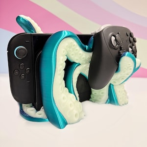 May include: A teal and off-white octopus-themed video game console stand. The stand holds a black game console and a black controller. The tentacles wrap around the console and controller, creating a unique and decorative design.