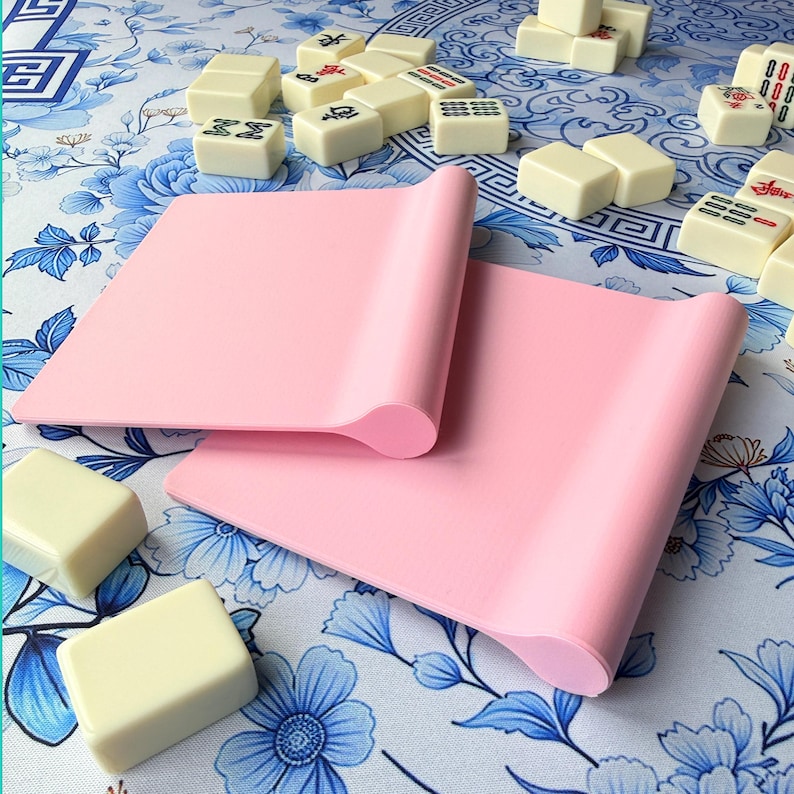 K&ouml;nnte beinhalten: Zwei rosa Silikonmatten mit gerollten Kanten werden auf einer Oberfl&auml;che mit floralem Muster gezeigt. Mehrere cremefarbene Mahjong-Steine mit schwarzen und roten Markierungen sind um die Matten herum verstreut. Der Hintergrund zeigt ein blau-wei&szlig;es Blumenmuster.