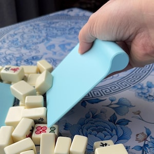 K&ouml;nnte beinhalten: Ein hellblauer Kunststoff-Pl&auml;ttchenschieber wird &uuml;ber einem Stapel elfenbeinfarbener Mahjong-Steine gehalten. Die Steine haben rote, gr&uuml;ne und schwarze Symbole. Der Hintergrund ist ein blau-wei&szlig;es Blumenmuster.
