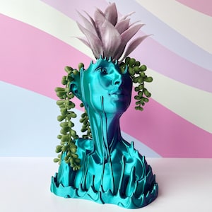 May include: A teal blue, 3D printed planter shaped like a woman's bust with a flowing, liquid-like design. The planter has a small opening at the top and is filled with green succulents and a purple flower.