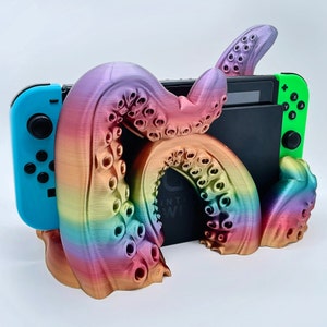May include: A rainbow-colored 3D printed octopus stand for a Nintendo Switch console. The stand has a textured, iridescent finish and features a detailed octopus design. The Switch console is partially visible in the stand.
