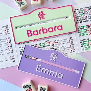 May include: Two personalized Mahjong tile holders, one light green with "Barbara" in pink, and one lavender with "Emma" in white. The holders have a Chinese character and are on a pink and white surface.