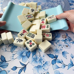 K&ouml;nnte beinhalten: Elfenbeinfarbene Mahjong-Steine mit roten, schwarzen und gr&uuml;nen Symbolen liegen auf einer Oberfl&auml;che mit floralem Muster. Ein hellblaues Tablett h&auml;lt einige der Steine. Die rechteckigen Spielsteine zeigen chinesische Schriftzeichen.