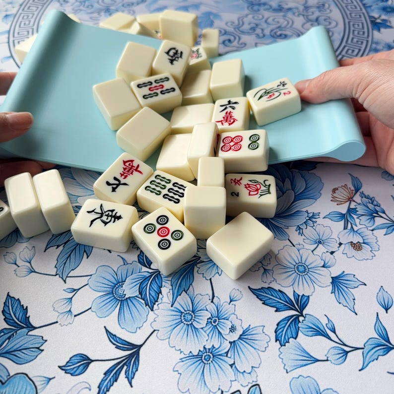 K&ouml;nnte beinhalten: Elfenbeinfarbene Mahjong-Steine mit roten, schwarzen und gr&uuml;nen Symbolen liegen auf einer Oberfl&auml;che mit floralem Muster. Ein hellblaues Tablett h&auml;lt einige der Steine. Die rechteckigen Spielsteine zeigen chinesische Schriftzeichen.