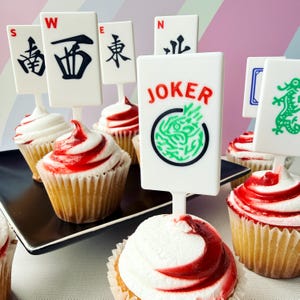 May include: Cupcakes with white frosting swirled with red, topped with white rectangular Mahjong tile toppers. The toppers have black Chinese characters and the word "JOKER" in red.