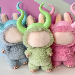 May include: Three plush monster dolls in blue, green, and pink. Each has a fluffy body, a round face, and large, curved horns. The dolls are standing in front of a colorful background.