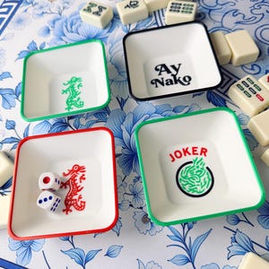 May include: Four white square dishes with colored rims and decorative elements. One dish has a green dragon, another has the text "Ay Nako", a third has a red dragon and dice, and the last has the word "JOKER" with a green design.