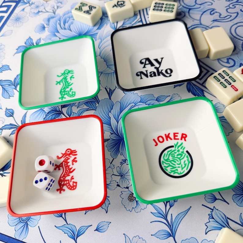 May include: Four white square dishes with colored rims and decorative elements. One dish has a green dragon, another has the text "Ay Nako", a third has a red dragon and dice, and the last has the word "JOKER" with a green design.