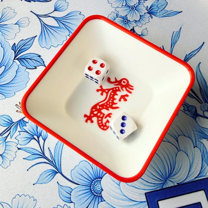 May include: A square, off-white ceramic bowl with a red rim, containing two dice and a red dragon design. The dice are white with red dots. The bowl sits on a blue and white floral patterned surface.