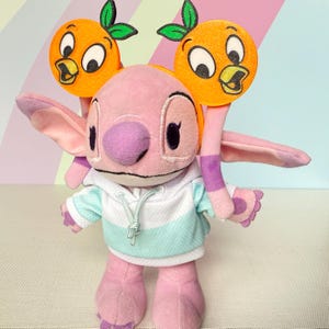 May include: A pink plush character wearing a white and light blue hooded sweatshirt. The character has large ears and is wearing a headband featuring two orange circular icons with green leaves and cartoon bird faces on top of the head.