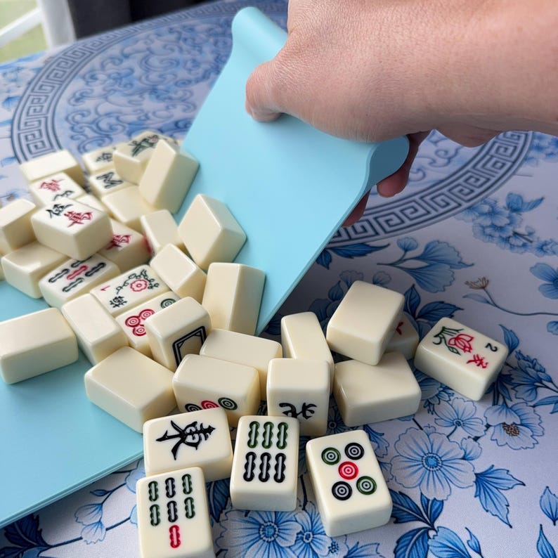 K&ouml;nnte beinhalten: Elfenbeinfarbene Mahjong-Steine ​​werden aus einem hellblauen Tablett gesch&uuml;ttet. Die Steine ​​zeigen rote, gr&uuml;ne und schwarze Symbole und Schriftzeichen. Die Spielsteine ​​liegen auf einer blau-wei&szlig; gebl&uuml;mten Oberfl&auml;che.
