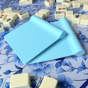 K&ouml;nnte beinhalten: Zwei hellblaue Silikonmatten mit gerollten Kanten, die auf einer blau-wei&szlig; gemusterten Oberfl&auml;che liegen. Verstreute elfenbeinfarbene Mahjong-Steine mit roten und schwarzen Symbolen sind um die Matten herum zu sehen.