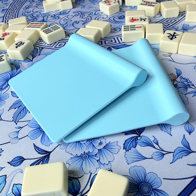 K&ouml;nnte beinhalten: Zwei hellblaue Silikonmatten mit gerollten Kanten, die auf einer blau-wei&szlig; gemusterten Oberfl&auml;che liegen. Verstreute elfenbeinfarbene Mahjong-Steine mit roten und schwarzen Symbolen sind um die Matten herum zu sehen.