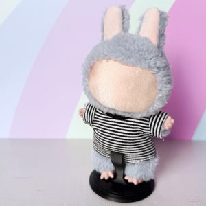 May include: A plush doll wearing a gray bunny hood with pink inner ears and a black and white striped shirt. The doll is standing on a black stand against a pastel background.