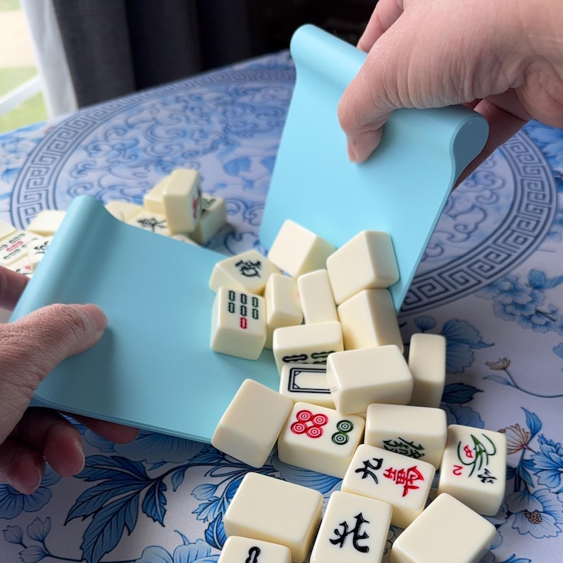 K&ouml;nnte beinhalten: Eine hellblaue Silikonmatte wird verwendet, um elfenbeinfarbene Mahjong-Steine auszusch&uuml;tten. Die Steine sind mit verschiedenen Symbolen und Zahlen in Rot und Schwarz markiert. Der Hintergrund zeigt ein blau-wei&szlig;es Blumenmuster.
