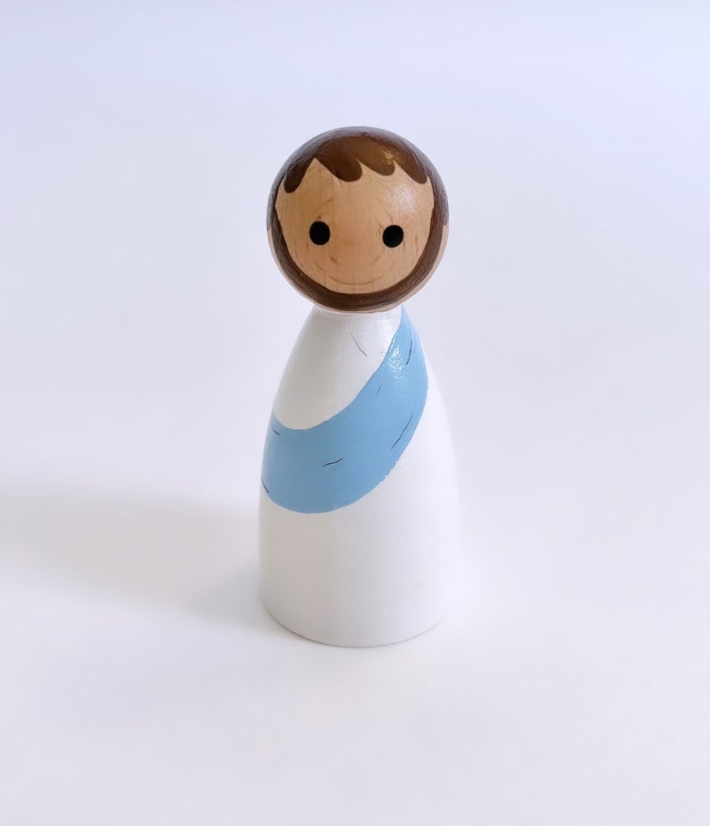 May include: A wooden doll with a white robe and a blue sash. The doll has a brown head with a painted face and brown hair.