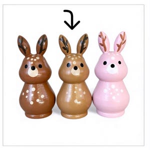 May include: Three wooden bunny figurines in brown, tan, and pink. Each bunny has a spotted pattern, black eyes, and brown antlers. The bunnies are painted with a glossy finish and have a rounded shape.